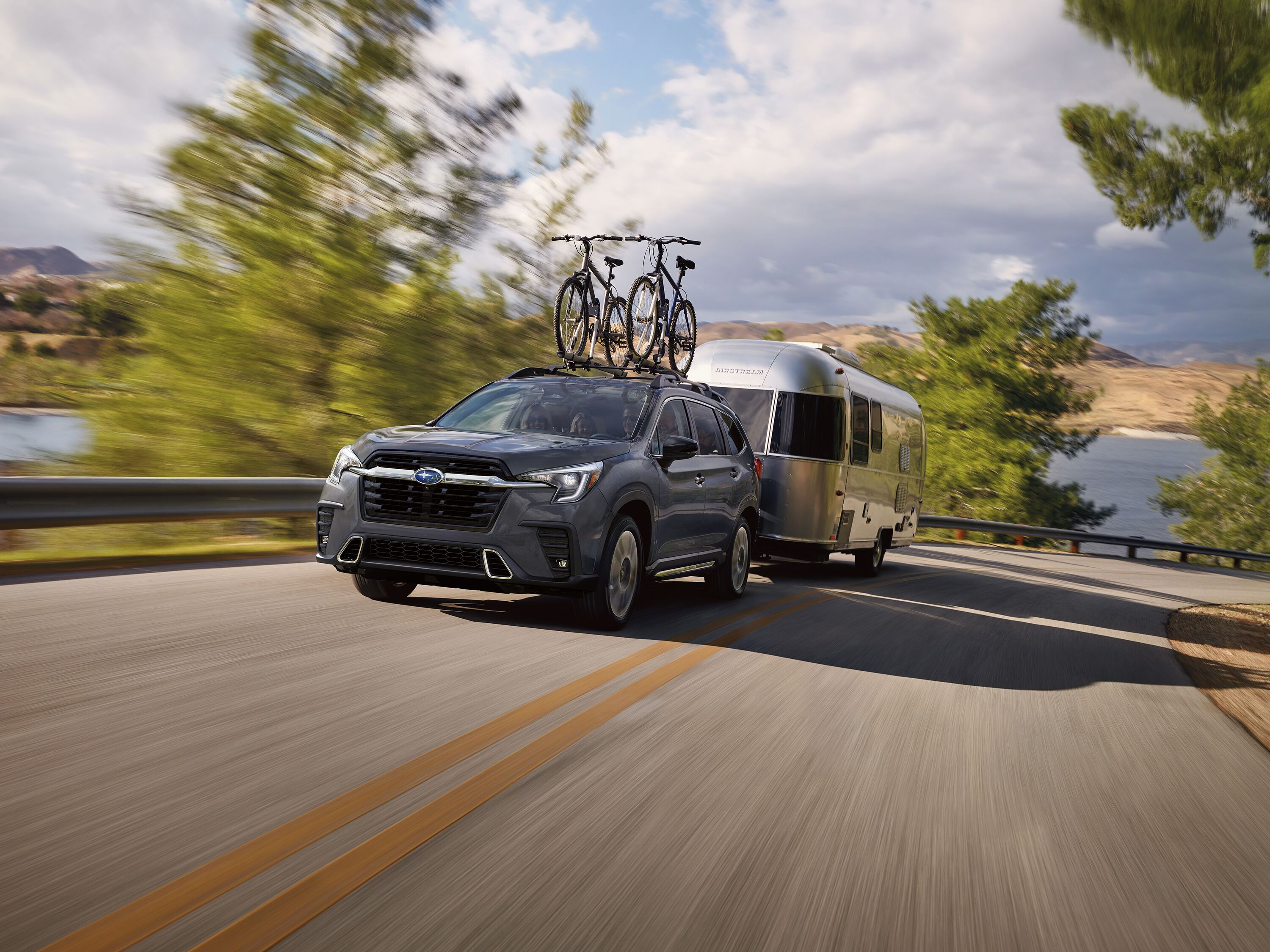 2027 Subaru Ascent Dark gray Subaru Ascent SUV with roof-mounted bikes towing a silver travel trailer along a coastal curve.