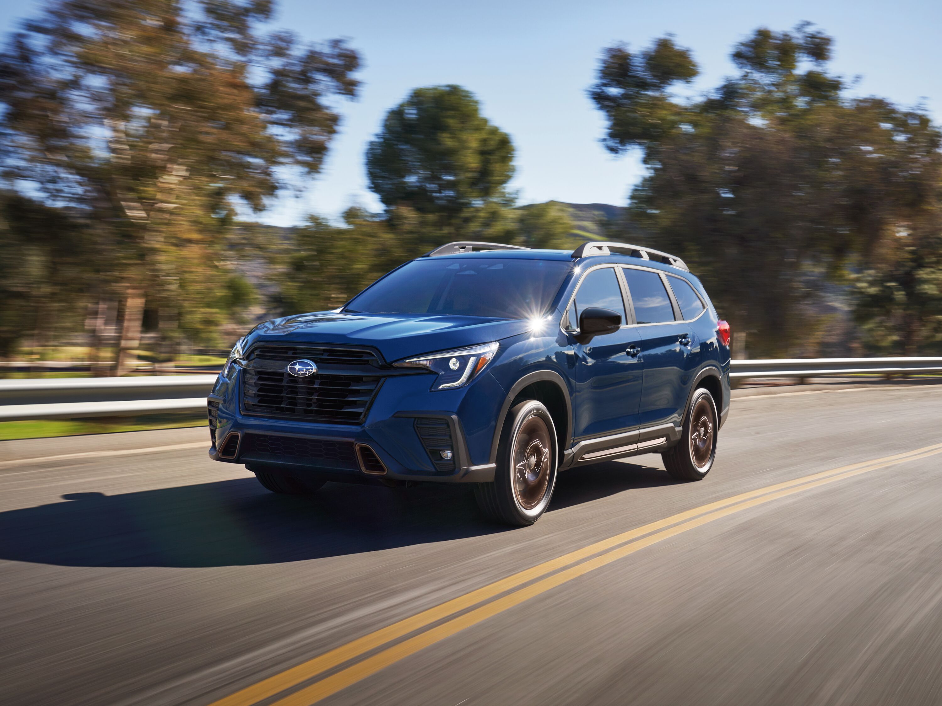 2027 Subaru Ascent Front three-quarter view of a Cosmic Blue Pearl Subaru Ascent 3-row SUV driving along a highway.