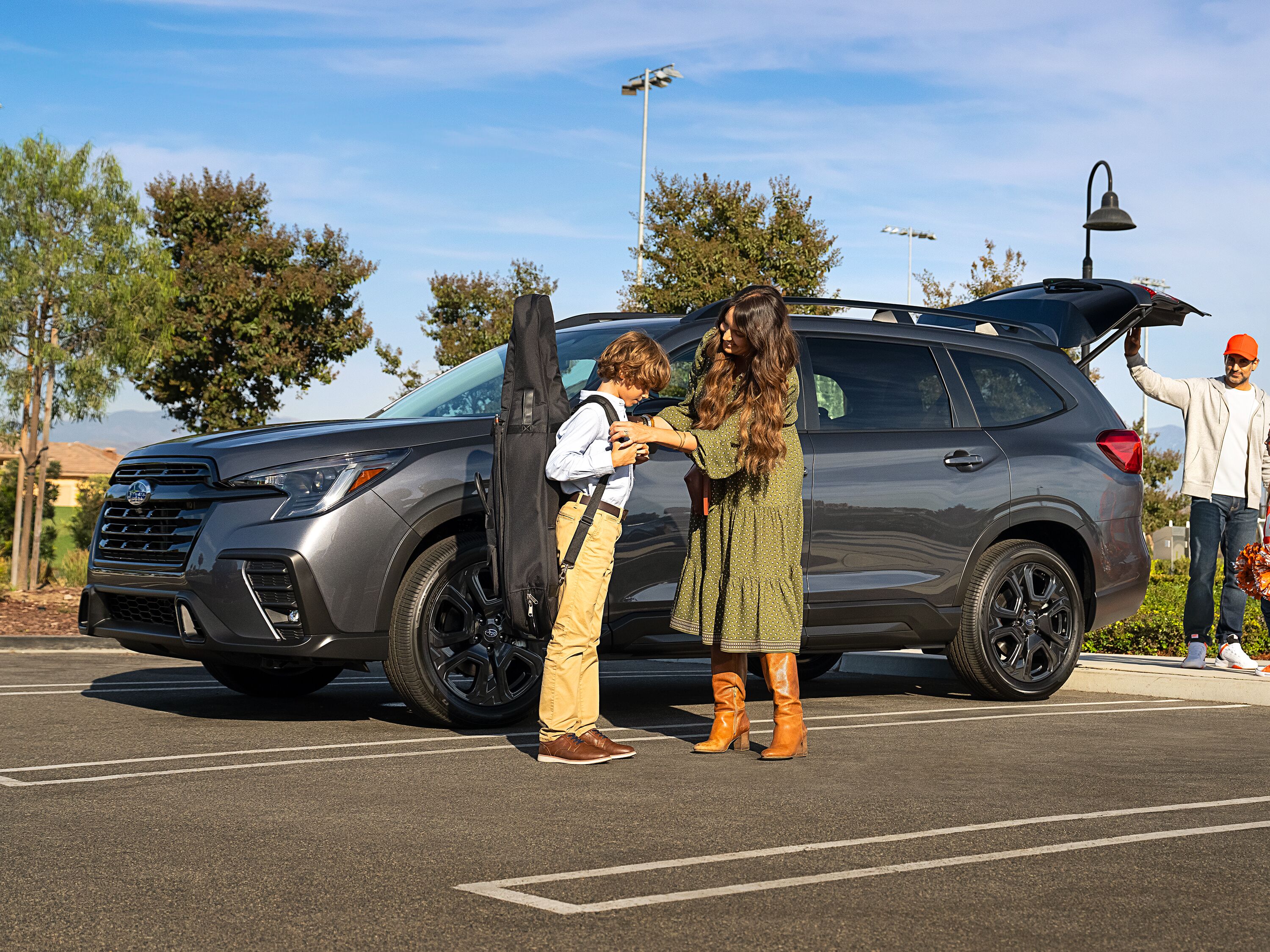 2027 Subaru Ascent Family packing musical instrument and luggage into the open rear cargo area of a gray Subaru Ascent SUV in a parking lot.