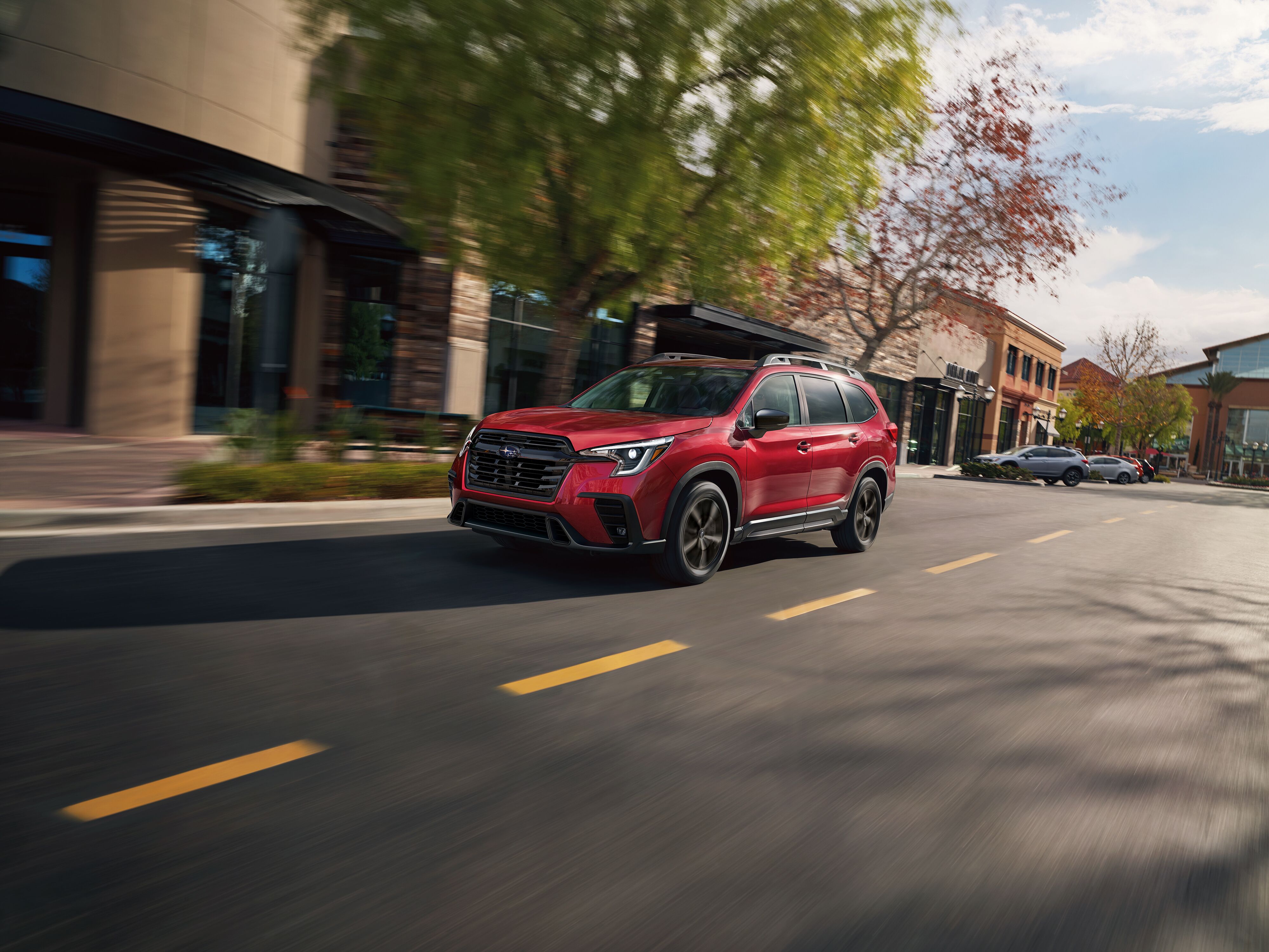 2027 Subaru Ascent Crimson Red Pearl Subaru Ascent SUV driving past shops and trees on a paved city street.