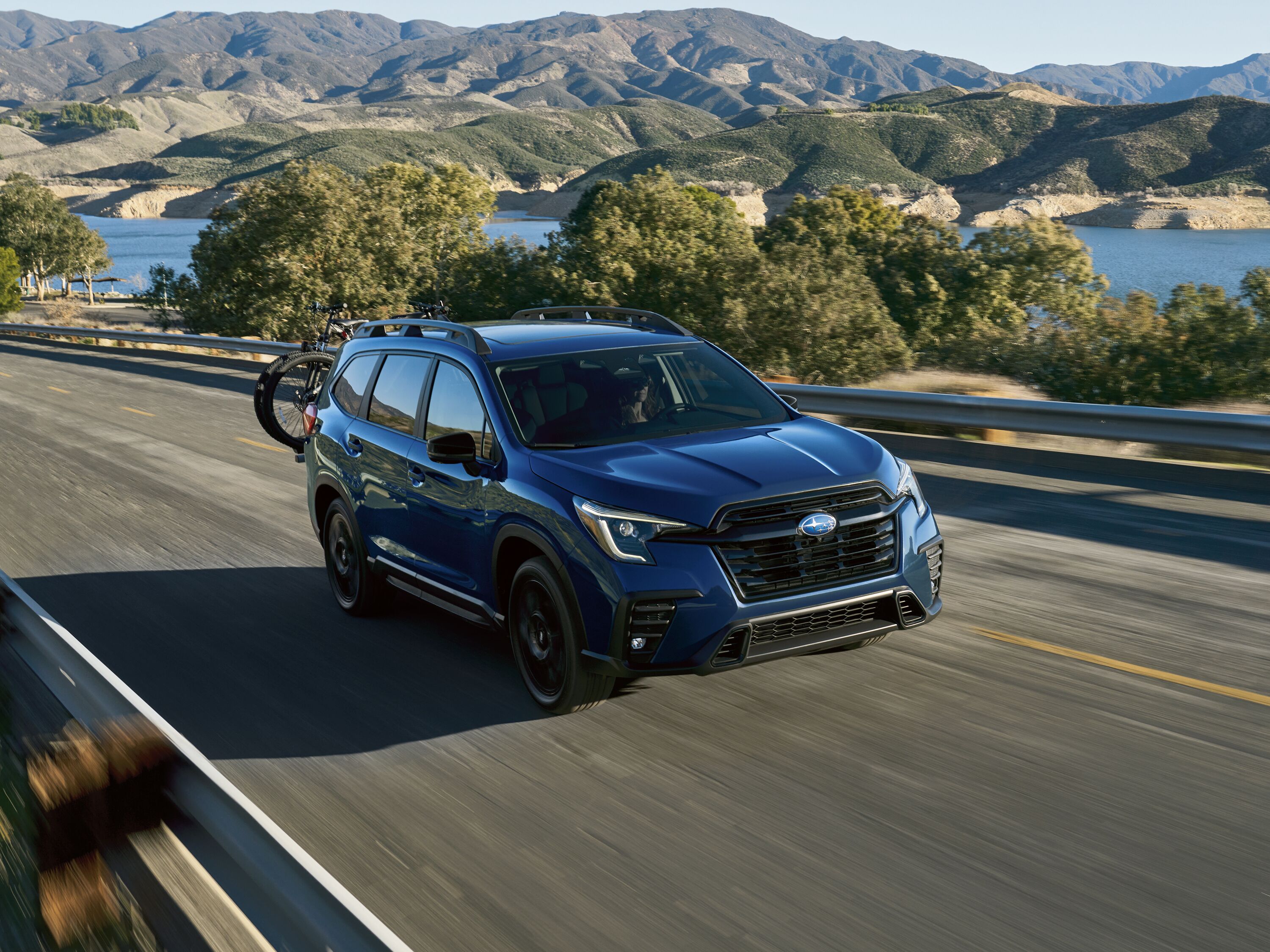 2027 Subaru Ascent Blue Subaru Ascent SUV equipped with a rear hitch bicycle rack driving along a scenic lakeside mountain road.