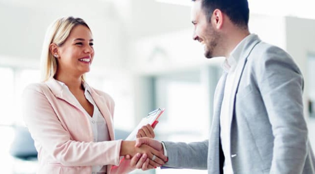A smiling woman in a light pink suit shakes hands with a smiling man in a light gray suit.