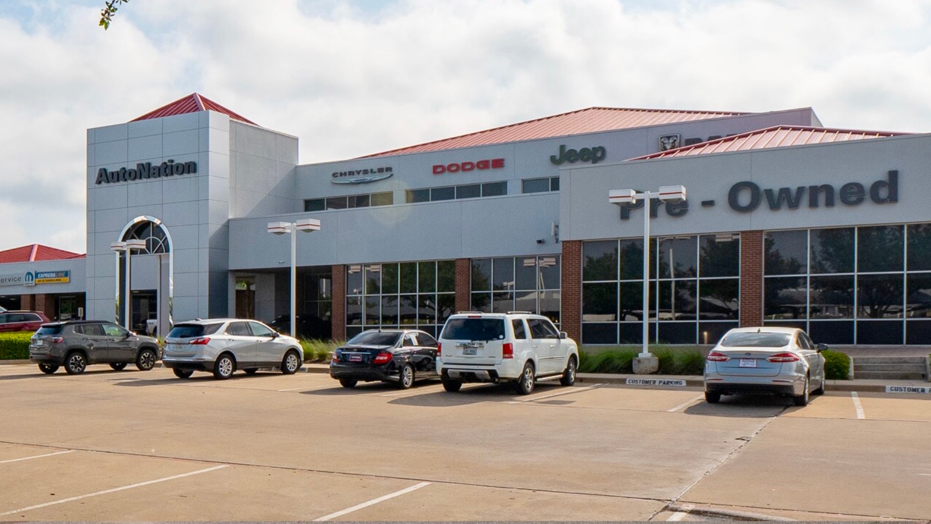 Exterior view of AutoNation Chrysler Dodge Jeep Ram North Fort Worth during the day. There is a cloudy sky and many vehicles parked near the grey and brick faced building. Trees and greenery are visible around the parking lot.
