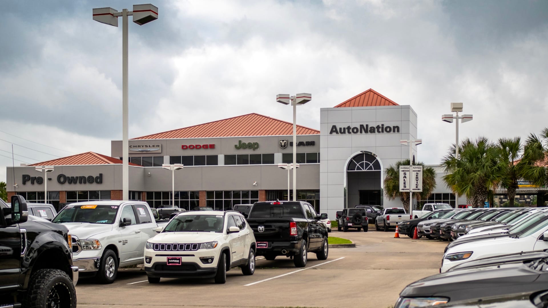 Exterior view of AutoNation Chrysler Dodge Jeep Ram Katy during the day. There is a cloudy sky and many vehicles parked near the grey and brick faced building. Trees and greenery are visible around the parking lot.