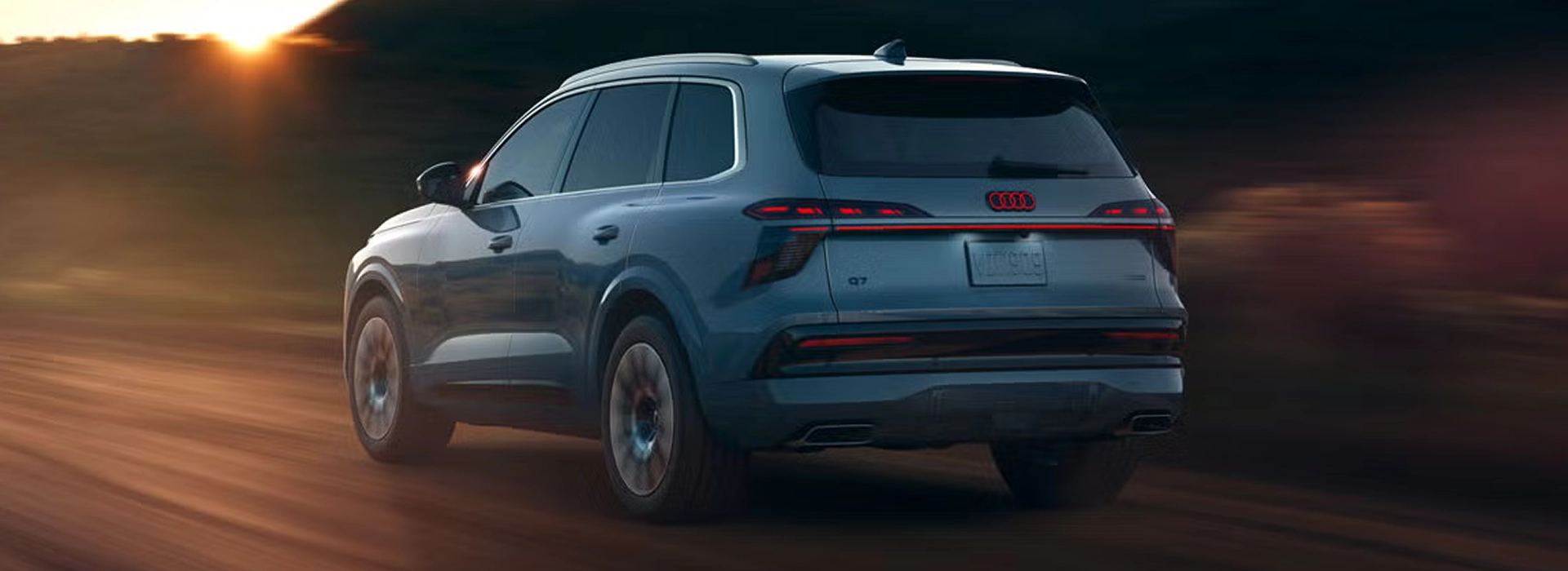 Blue-gray Audi Q7 driving away on a dirt road at sunset, rear three-quarter view