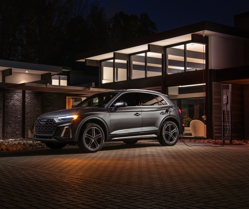 2021-Audi-Q5-Hybrid-SUV-L10.png silver Audi Q5e parked in front of a night-lit modern home, plugged into a charging station