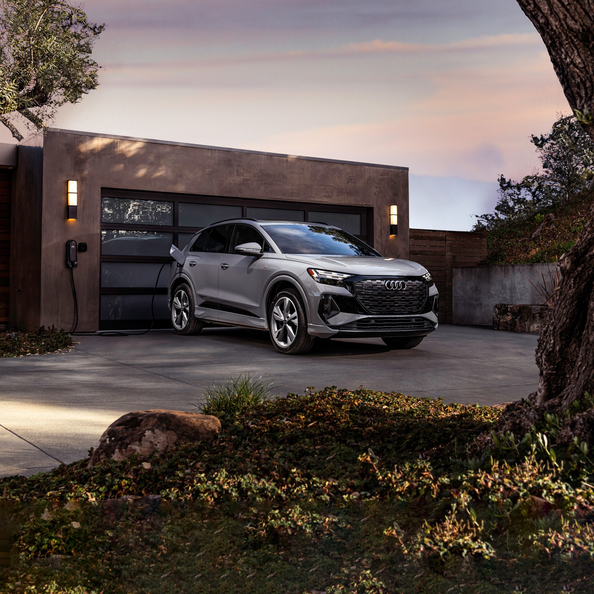 silver Audi Q4 e-tron parked in front of a modern garage, plugged in to a charging station