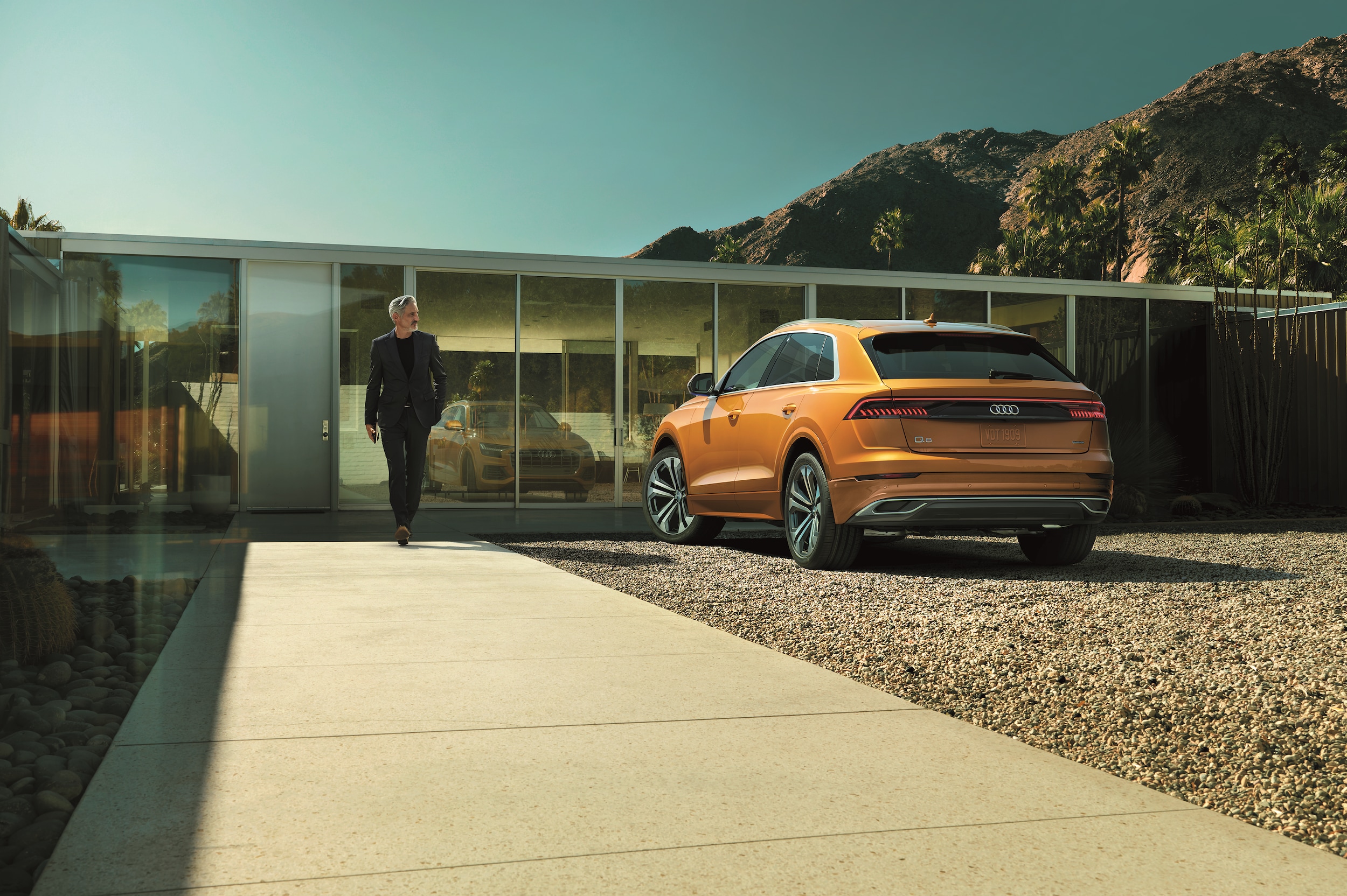 2022-Audi-Q8-PremiumPlus-WheelAddOnSUV-L01.png orange Audi Q8 parked in front of a modern class and cement home with the driver walking away