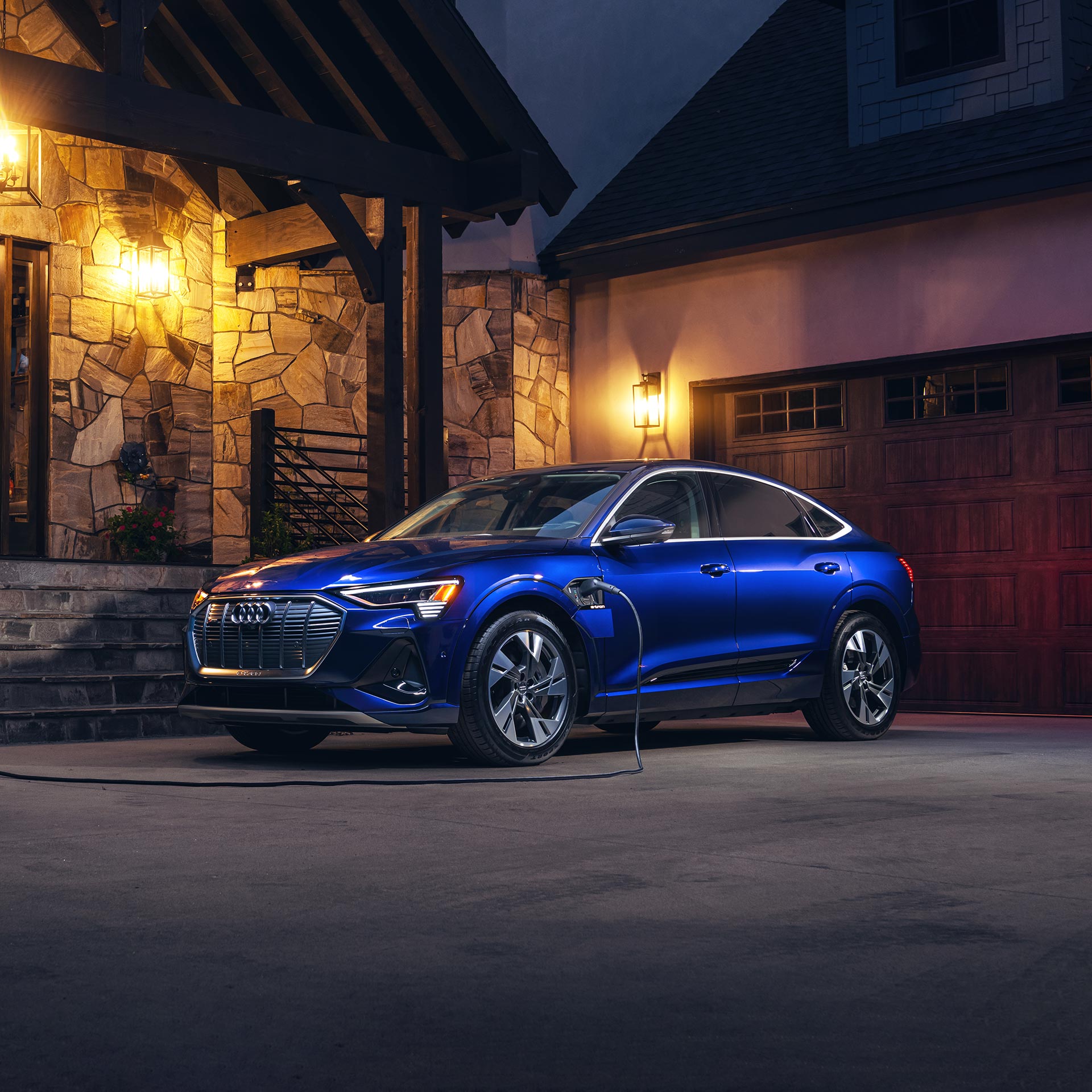 blue Audi e-tron parked in front of a modern garage and plugged in, charging