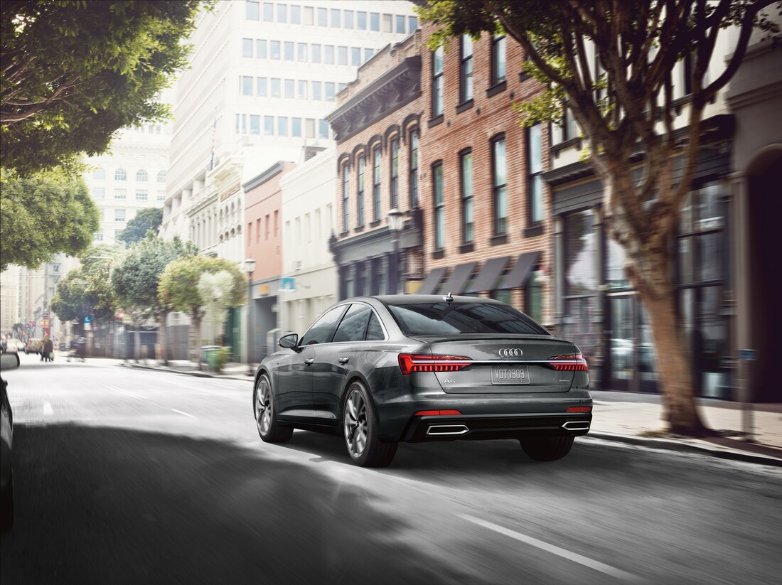 2022-Audi-A6-55TSI-PremiumPlus-WheelAddOn-Sedan-L01.png silver Audi A6 sedan driving away down a tree-lined street with brick buildings