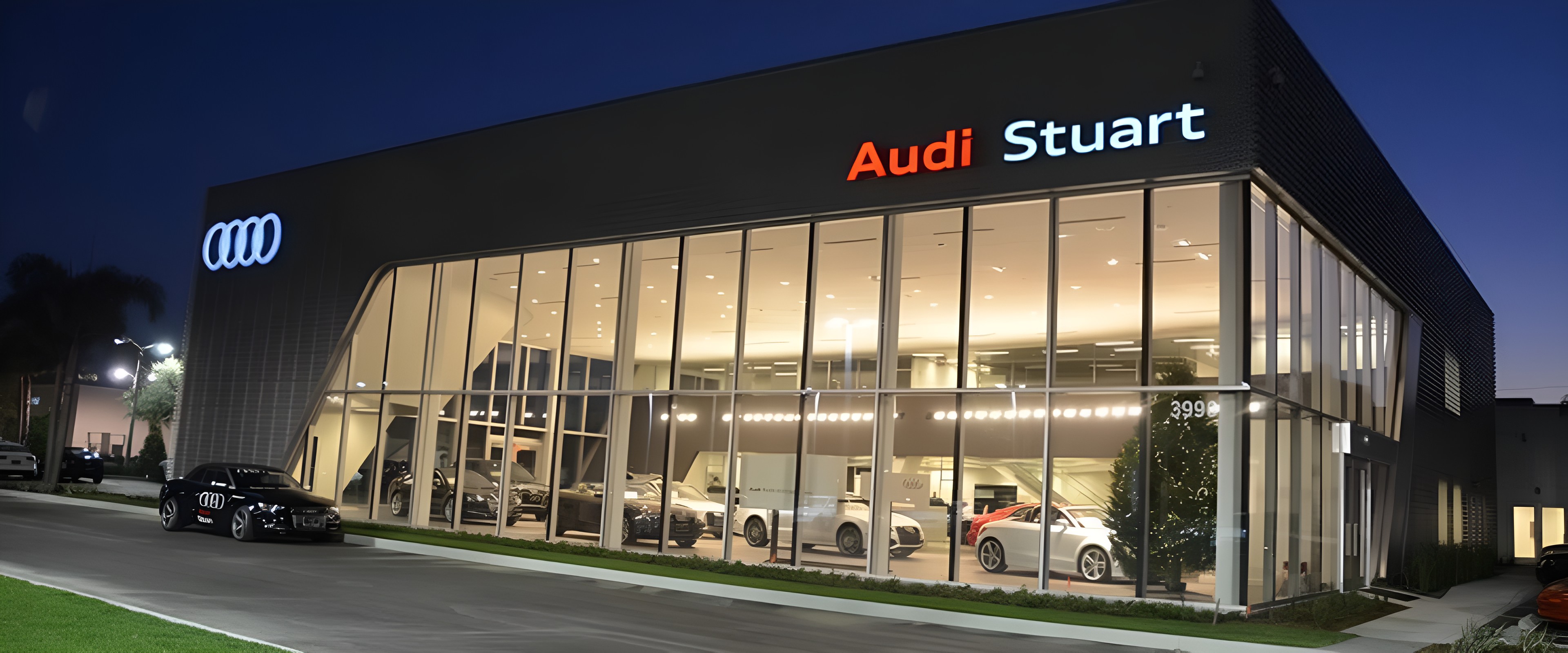 The Audi Stuart dealership at dusk, its glass showroom lit up beneath the Audi Stuart sign, with a black Audi parked out front