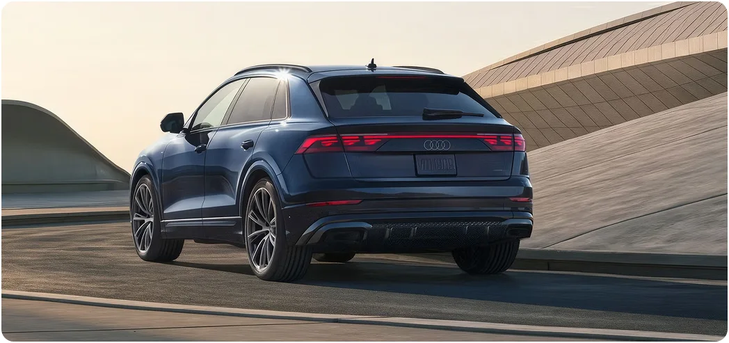 Blue Audi Q8 with black optic trim driving past a curved concrete structure, rear three-quarter view