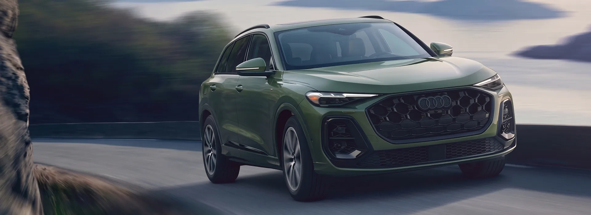 Dark green 2026 Audi Q5 driving a coastal mountain road, seen from the front three-quarter angle