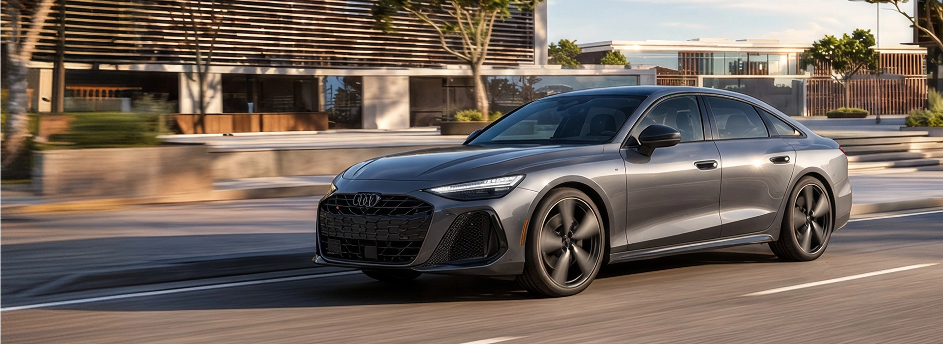 Grey 2027 Audi A6 Sedan driving past a modern building, seen from the front three-quarter angle