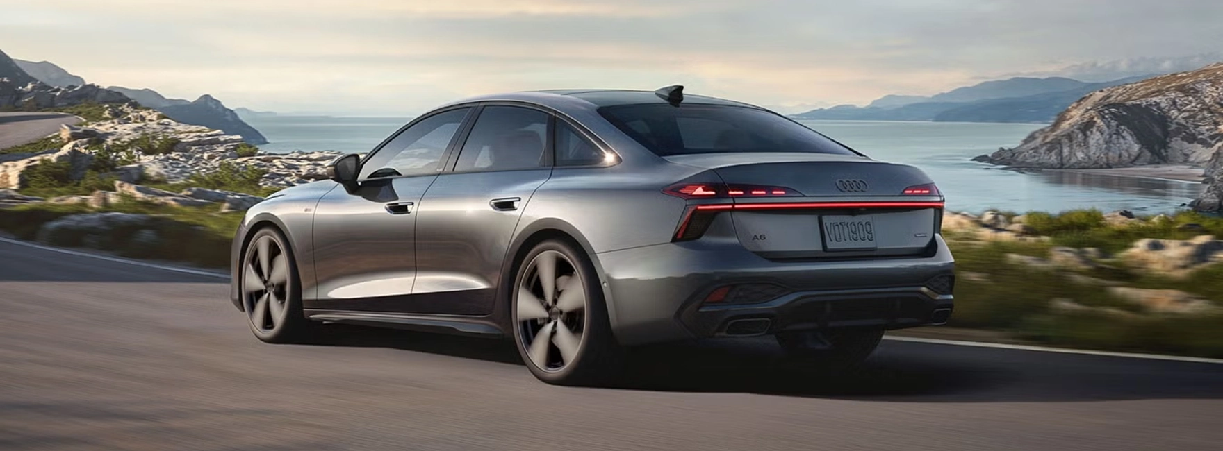 Grey Audi A6 Sedan driving along a coastal road, seen from the rear three-quarter angle