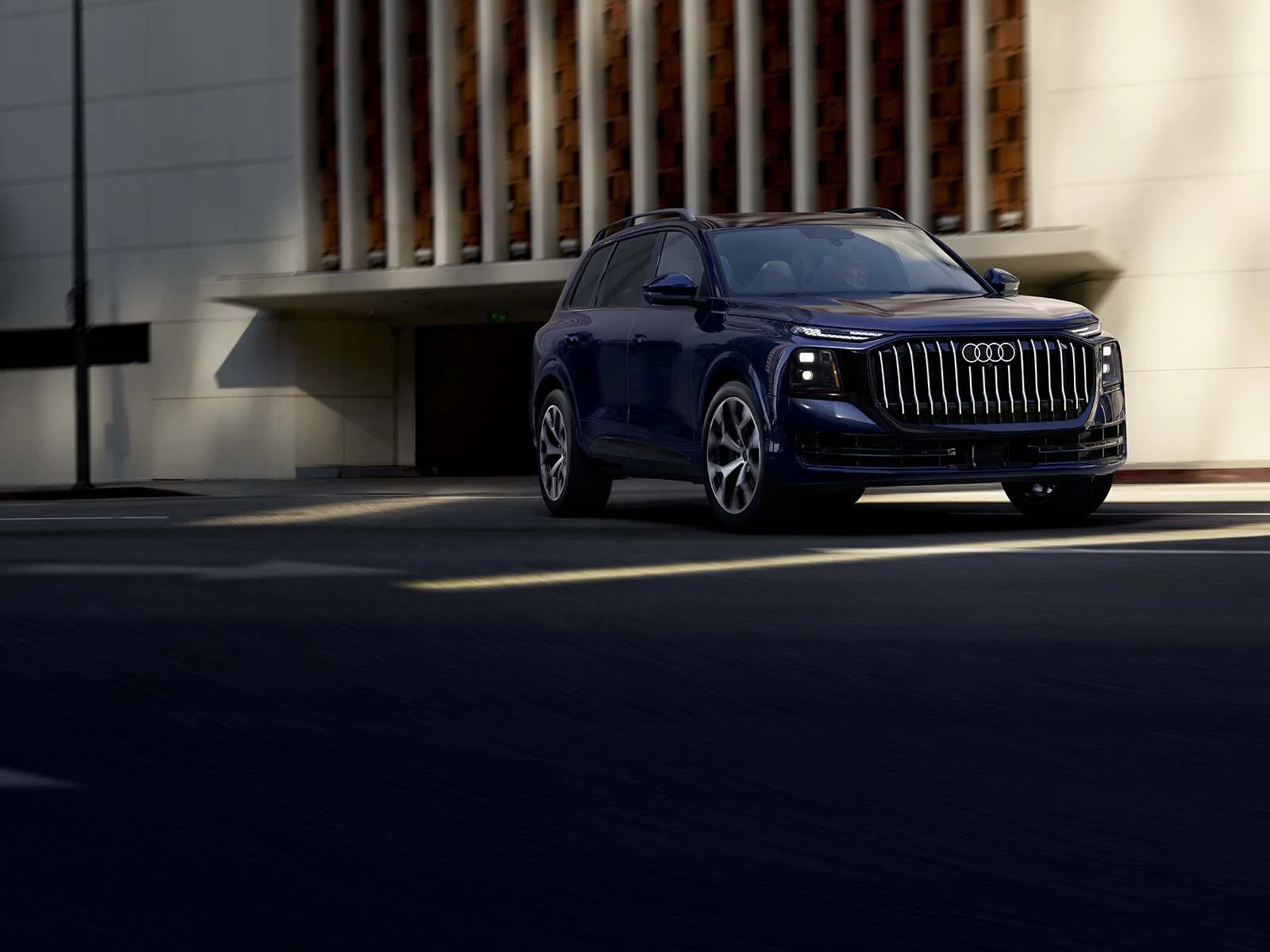 2027 Audi Q9 driving past a sunlit building, front three-quarter view