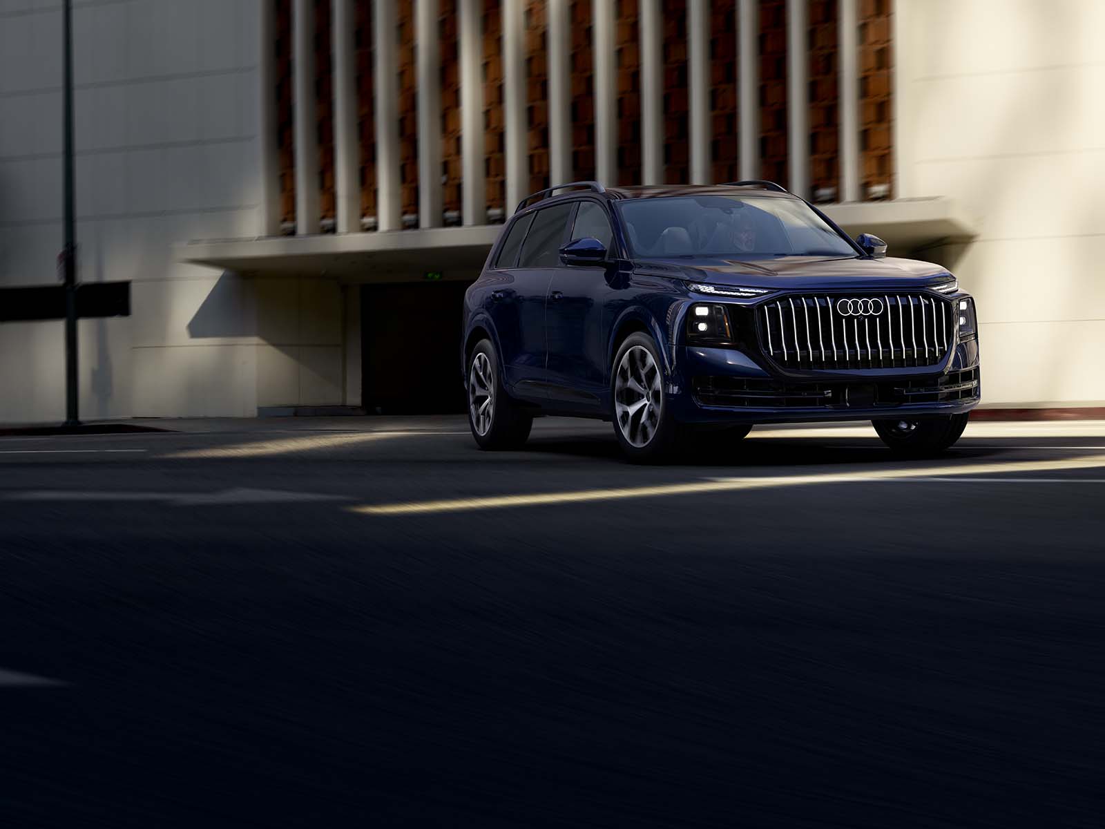 2027 Audi Q9 driving past a sunlit building, front three-quarter view