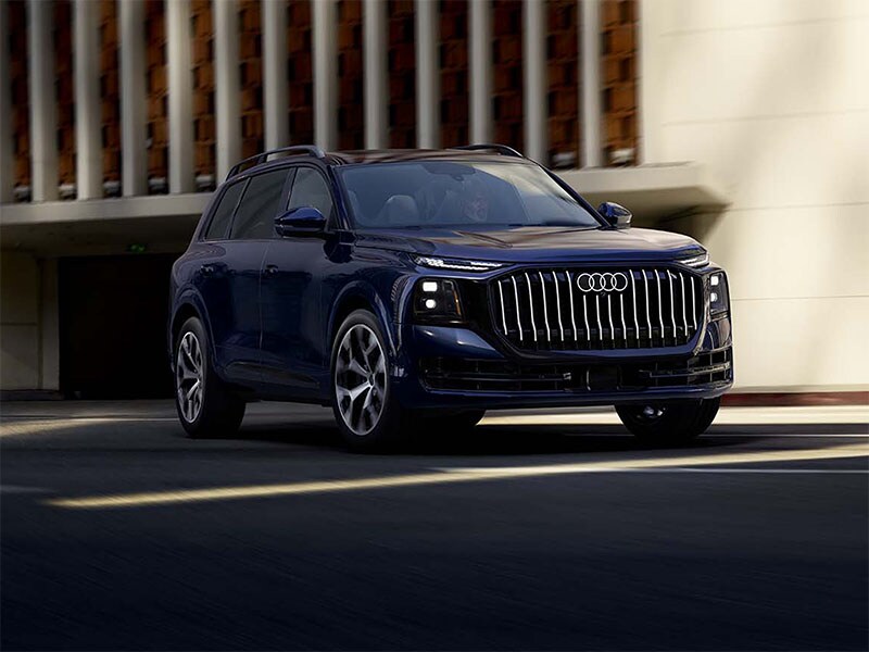 2027 Audi Q9 driving past a sunlit building, close front three-quarter view