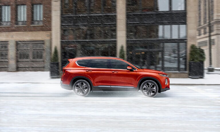2020 Hyundai SantaFe in orande side view from the passanger side driving in the snow throughout a town