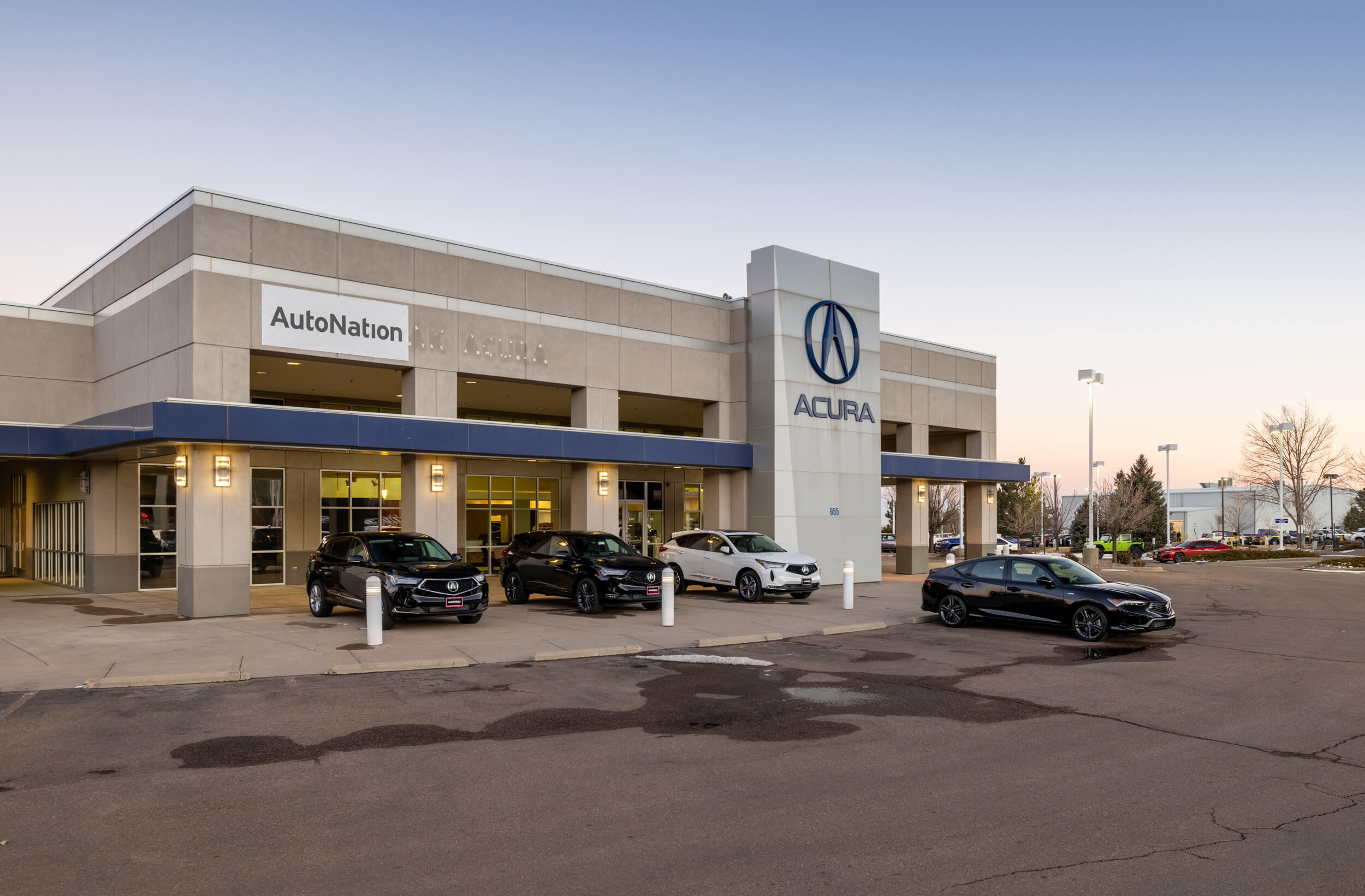 Exterior view of front of AutoNation Acura Colorado Springs with some cars parked in front on display. Clear sky in what appears to be the morning