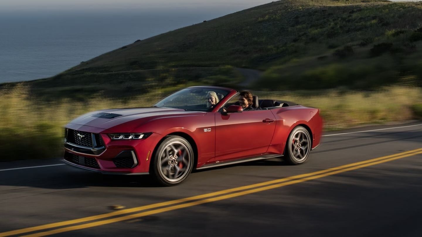 Exterior shot of a red Ford Mustang convertible driving along a coastline highway.