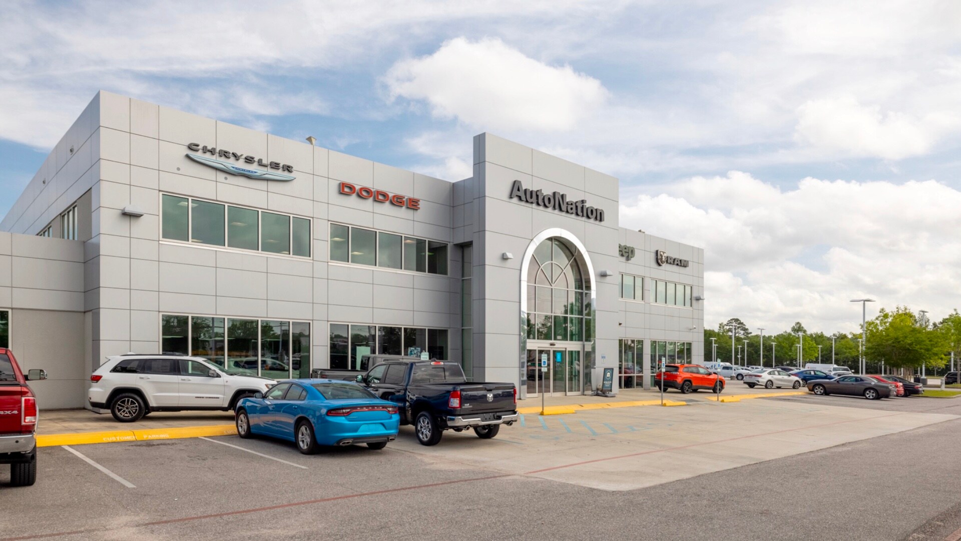 Exterior view of AutoNation Chrysler Dodge Jeep Ram Mobile during the day. There is a cloudy sky and many vehicles parked near the grey building. Trees and greenery are visible around the parking lot.