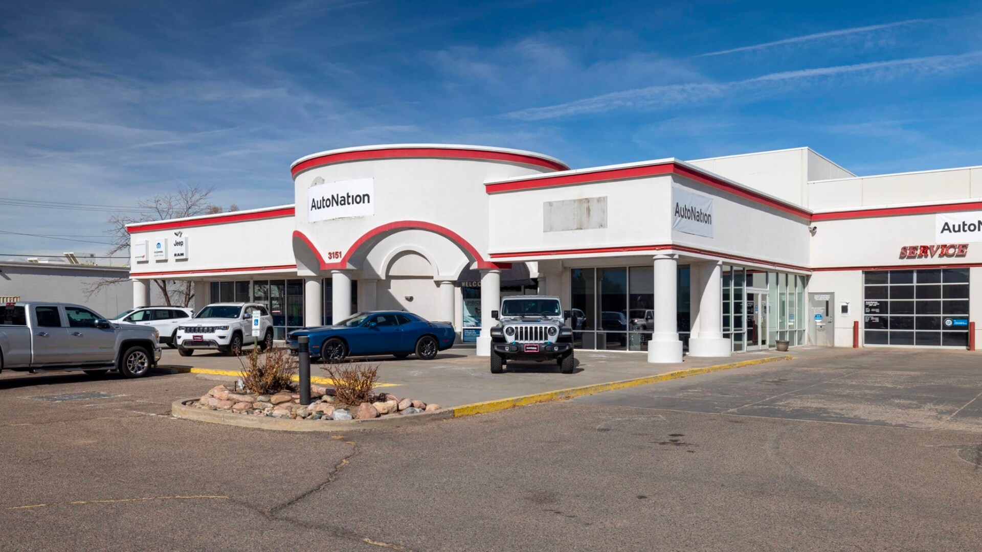 AutoNation Chrysler Dodge Jeep Ram Canon City is your Canon City, CO home for all your Dodge, Jeep, Chrysler, Ram financing, purchasing, and service needs Exterior view of AutoNation Chrysler Dodge Jeep Ram Canon City. There is a cloudy sky and many vehicles parked near the white building with a red stripe across the top. Trees and greenery are visible around the parking lot and surrounding area.