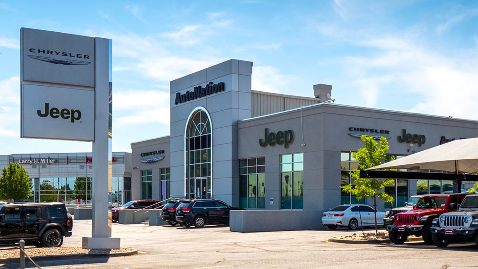 Exterior view of Autonation Chrysler Jeep Arapahoe during a clear sunny day. Many vehicles parked near the grey building. Trees and greenery are visible around the parking lot and surrounding area.