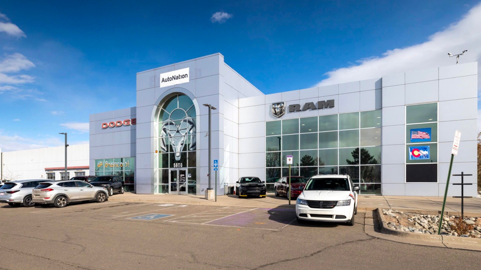 AutoNation Dodge Ram Broadway is your Littleton, CO home for all your Dodge, Ram financing, purchasing, and service needs Exterior view of AutoNation Dodge Ram Broadway. There is a partially cloudy blue sky and many vehicles parked near the grey building. Trees and greenery are visible around the parking lot and surrounding area.