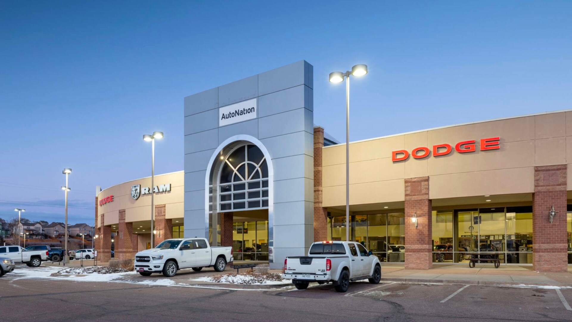 Exterior view of AutoNation Dodge Ram Colorado Springs. There is a blue sky and many vehicles parked near the grey building. Small amounts of snow can be sean near the building.
