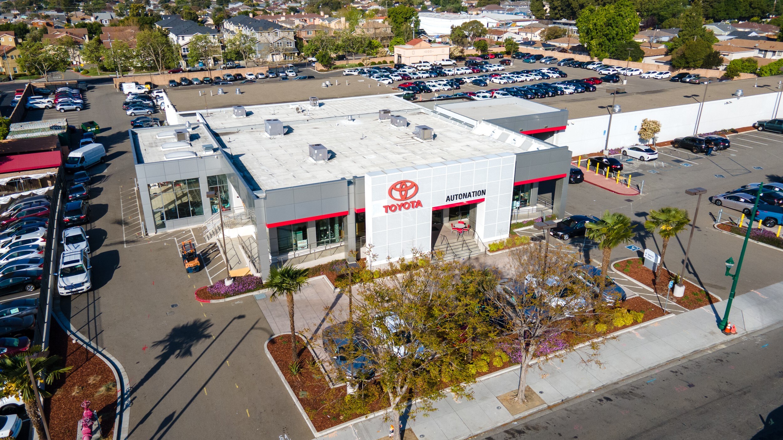 Overhead exterior view of AutoNation Toyota Hayward. The building is grey and white and has some large windows. Many vehicles can be seen parked near the building along with trees and homes in the background.