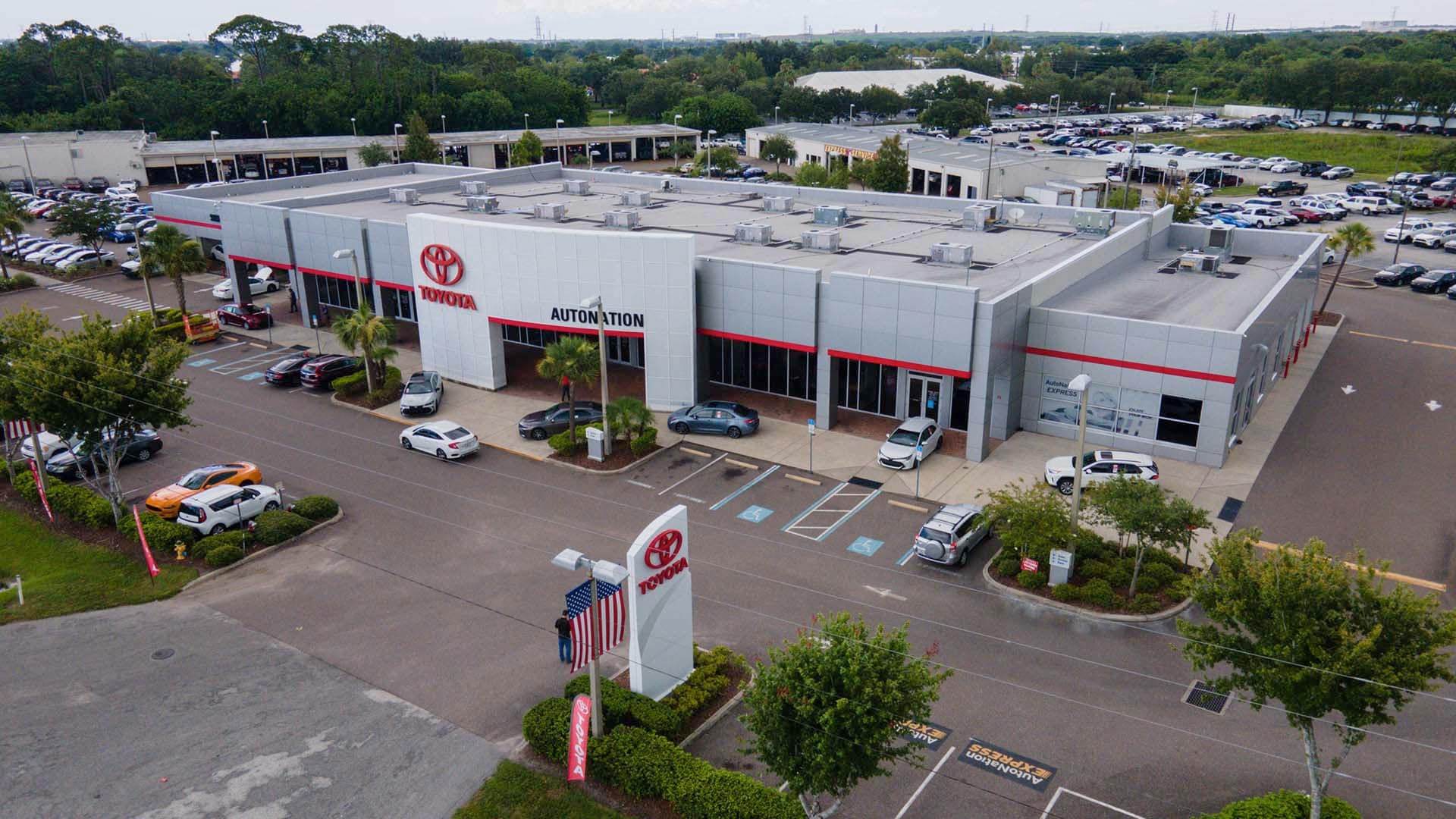 Overhead exterior view of AutoNation Toyota Pinellas Park. The building is grey and white and has some large windows. Many vehicles are seen parked in rows near the building, which also has some trees nearby.