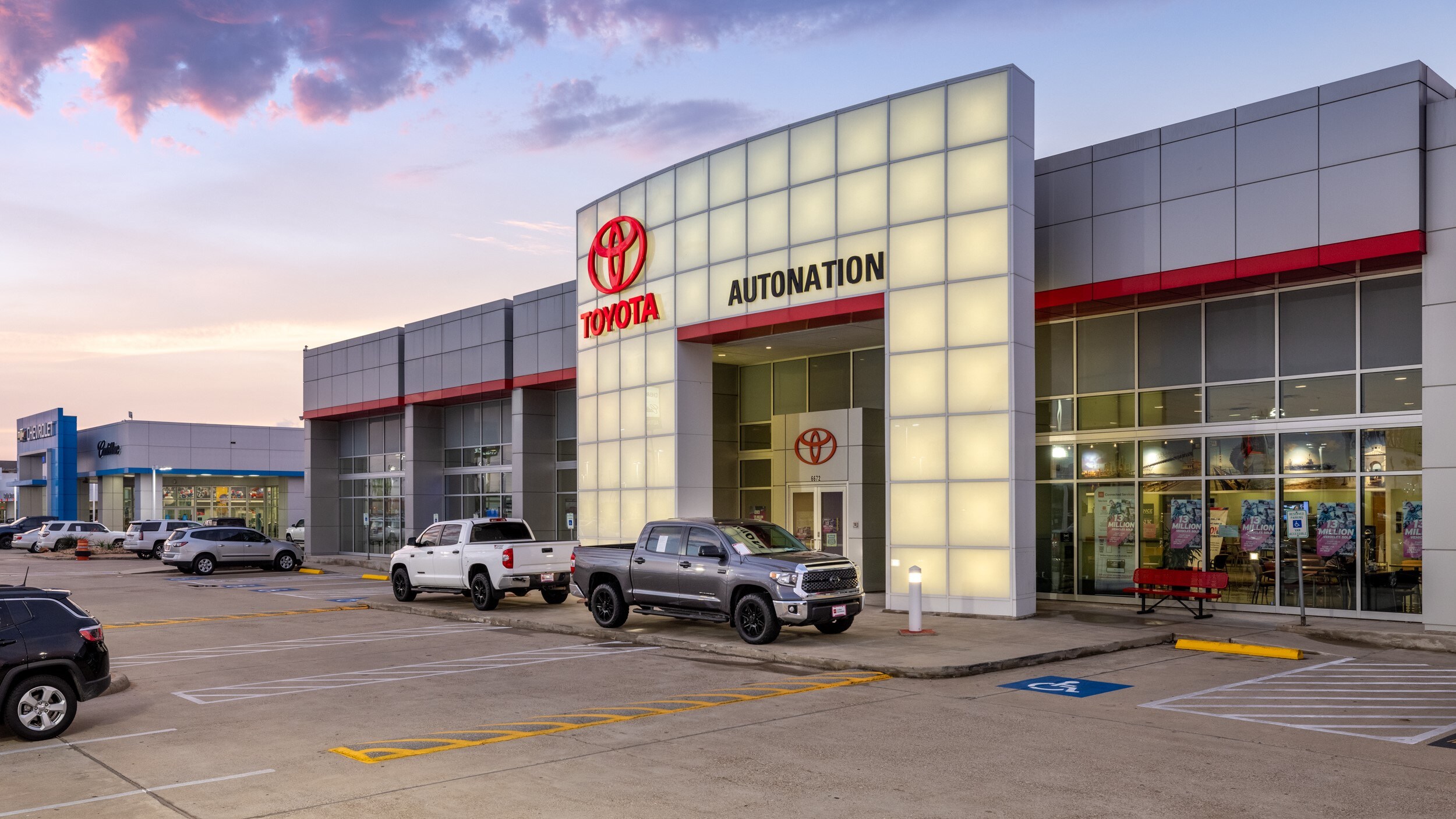 Exterior view of AutoNation Toyota Corpus Christi, with a partly cloudy blue sky. The building is grey, and part of the face is illuminated. Several vehicles can be seen parked near the vehicle.