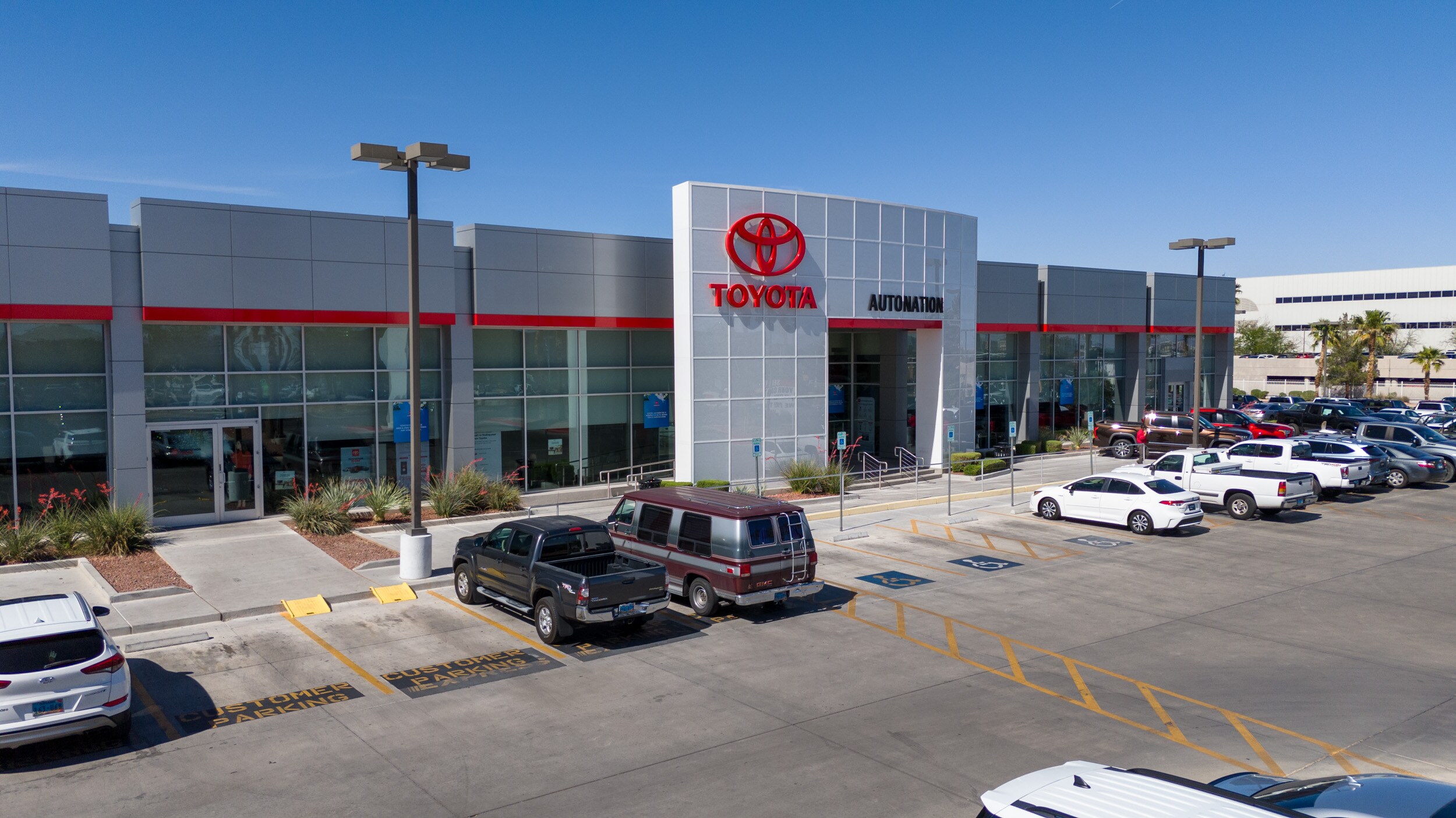 AutoNation Toyota Las Vegas is your Las Vegas, NV home for all your Toyota financing, purchasing, and service needs Overhead exterior view of AutoNation Toyota Las Vegas. The building is grey and white and has some large windows. Many vehicles can be seen parked near the building and on the roof. Mountains are visible in the distance.