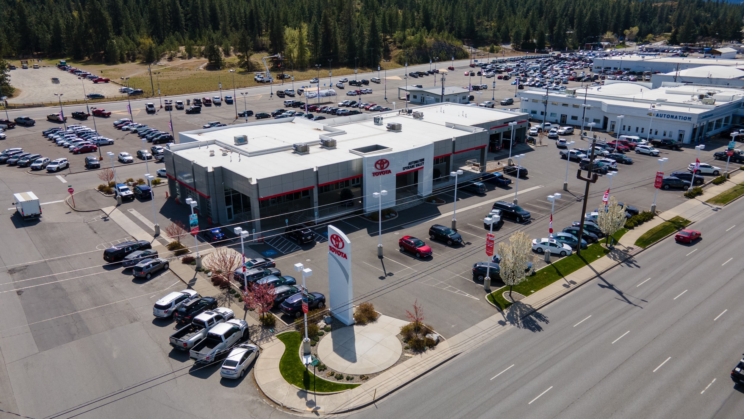 AutoNation Toyota Spokane Valley is your Spokane Valley, WA home for all your Toyota financing, purchasing, and service needs Overhead exterior view of AutoNation Toyota Spokane Valley. The building is grey and white and has some large windows. Many vehicles are seen parked in rows near the building, which also has many trees nearby in the background.