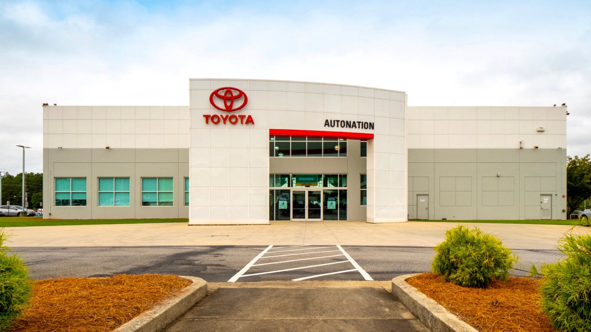 Exterior view of AutoNation Toyota Thornton Road on a partly cloudy day. The building is grey and white and has some large windows. Several vehicles can be seen near the building, which also has trees near nearby.