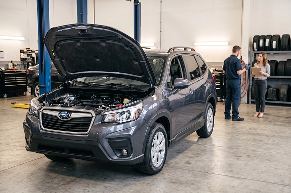A gray Subaru Forester with its hood raised in the Baldwin Subaru service bay while an advisor reviews a checklist with a technician