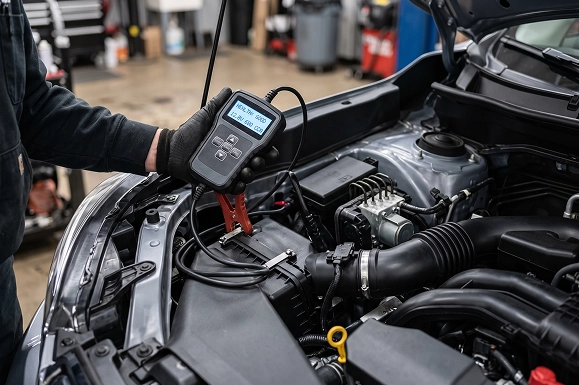A Baldwin Subaru technician holding a digital battery tester, its display lit, above the engine bay of a Subaru
