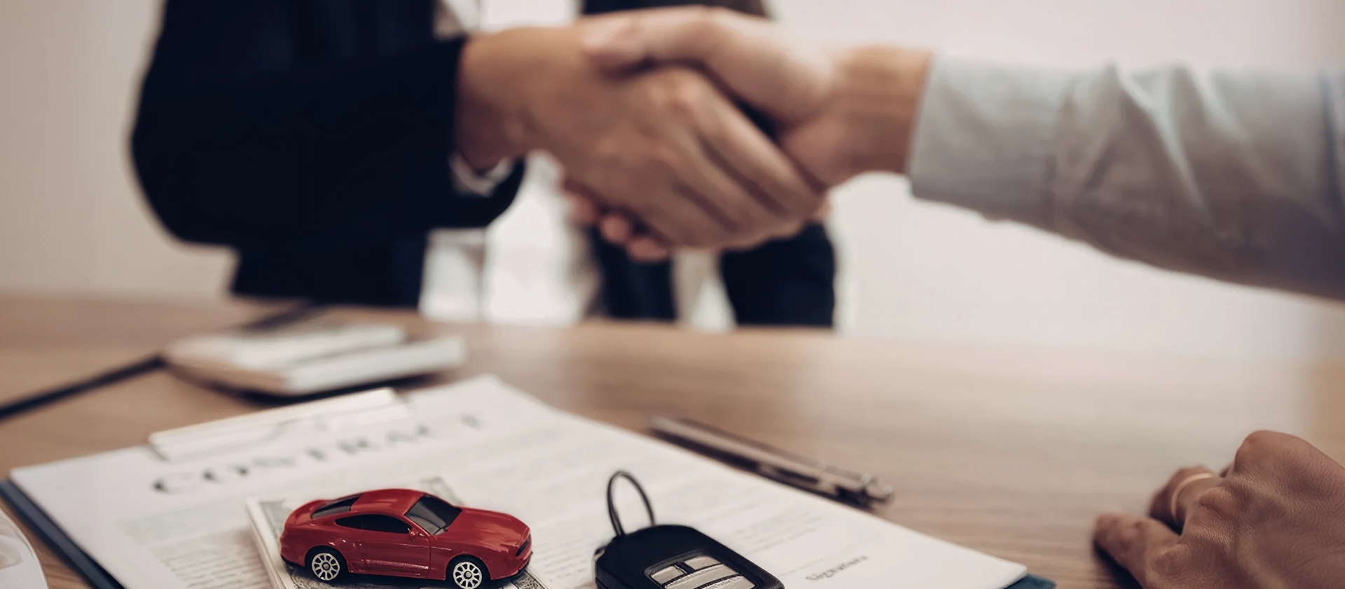 Two people shaking hands over a desk holding a signed contract, car keys and a red toy car