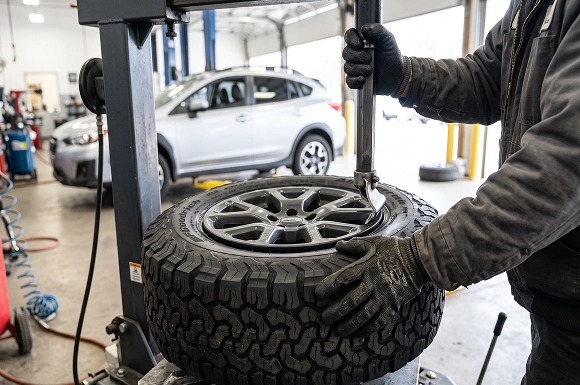 A close-up of a gloved Baldwin Subaru technician lowering the arm of a tire machine onto an all-terrain tire and alloy wheel
