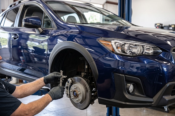 A close-up of a Baldwin Subaru technician's gloved hands fitting a caliper over the front brake rotor of a dark blue Subaru Crosstrek