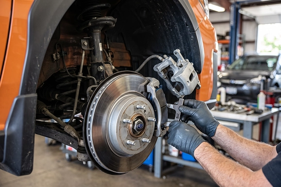 A close-up of a gloved Baldwin Subaru technician holding a brake pad beside the exposed rotor and caliper of an orange Subaru