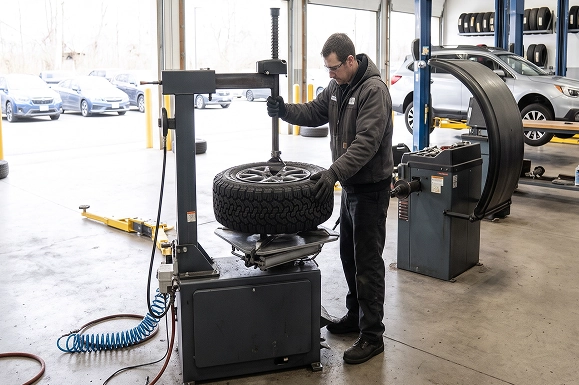 A Baldwin Subaru technician operating the tire machine to mount a wheel, with the service drive and parked Subarus visible through the bay door behind him
