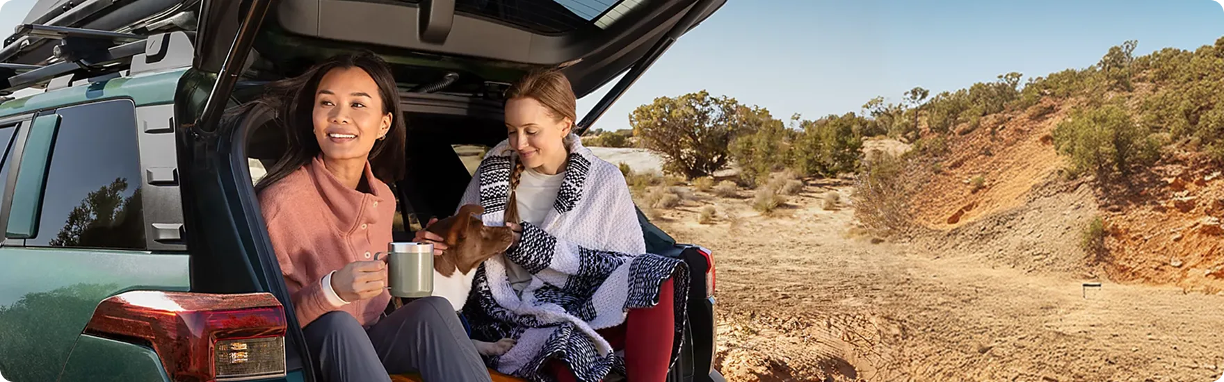 Two people sitting in the open rear gate of a Subaru Outback with gear beside them