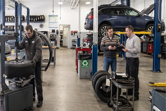 A Baldwin Subaru technician working at a tire machine while a service advisor reviews a tablet with a customer, a Subaru Outback raised on a lift behind them
