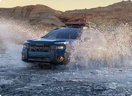 A blue Subaru Outback sending up spray as it fords shallow water at sunset
