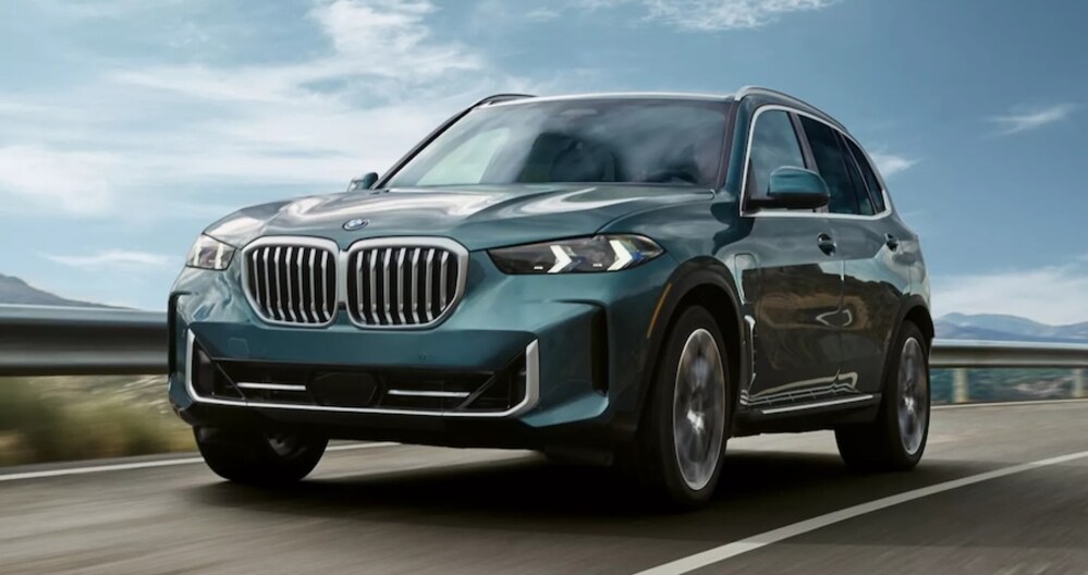 A gray-blue 2026 BMW X5 SUV driving down the road with mountains in the background