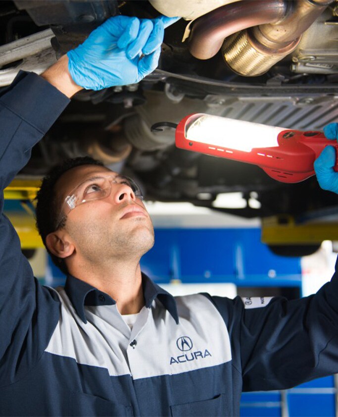 Acura Maintenance At Bobby Rahal Acura in Mechanicsburg, PA | Mechanic in Acura uniform is inspection the bottom of a vehicle while using a red light stick and safety glasses