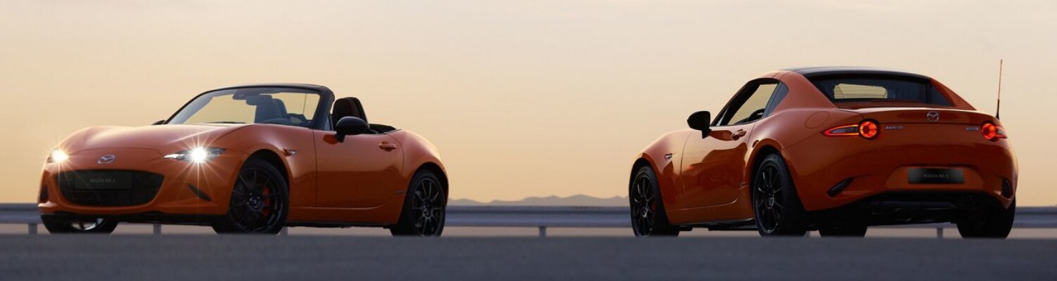 2019 Mazda Miata MX-5 in Colorado Springs Bob Penkhus Dealerships.png An orange 2019 Mazda Miata MX-5 and 2019 Mazda MX-5 RF parked in an open lot in front of a vibrant orange sunset
