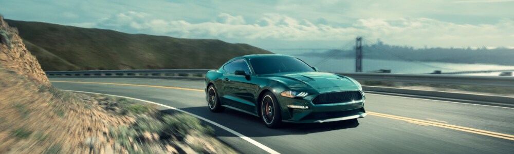 New 2019 Ford Mustang near Seattle at Bowen Scarff Ford in Kent, WA.png Zoomed out front passenger side angle of a dark green 2019 Ford Mustang speeding down a cliff side curved road overlooking a bridge crossing a body of water